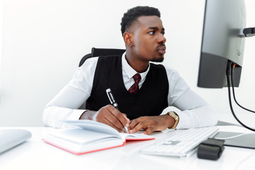 A man in formal attire writes in a notebook while looking at a computer screen