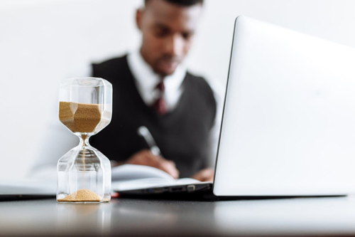 A man sitting at a desk with a laptop and an hourglass filled with sand.